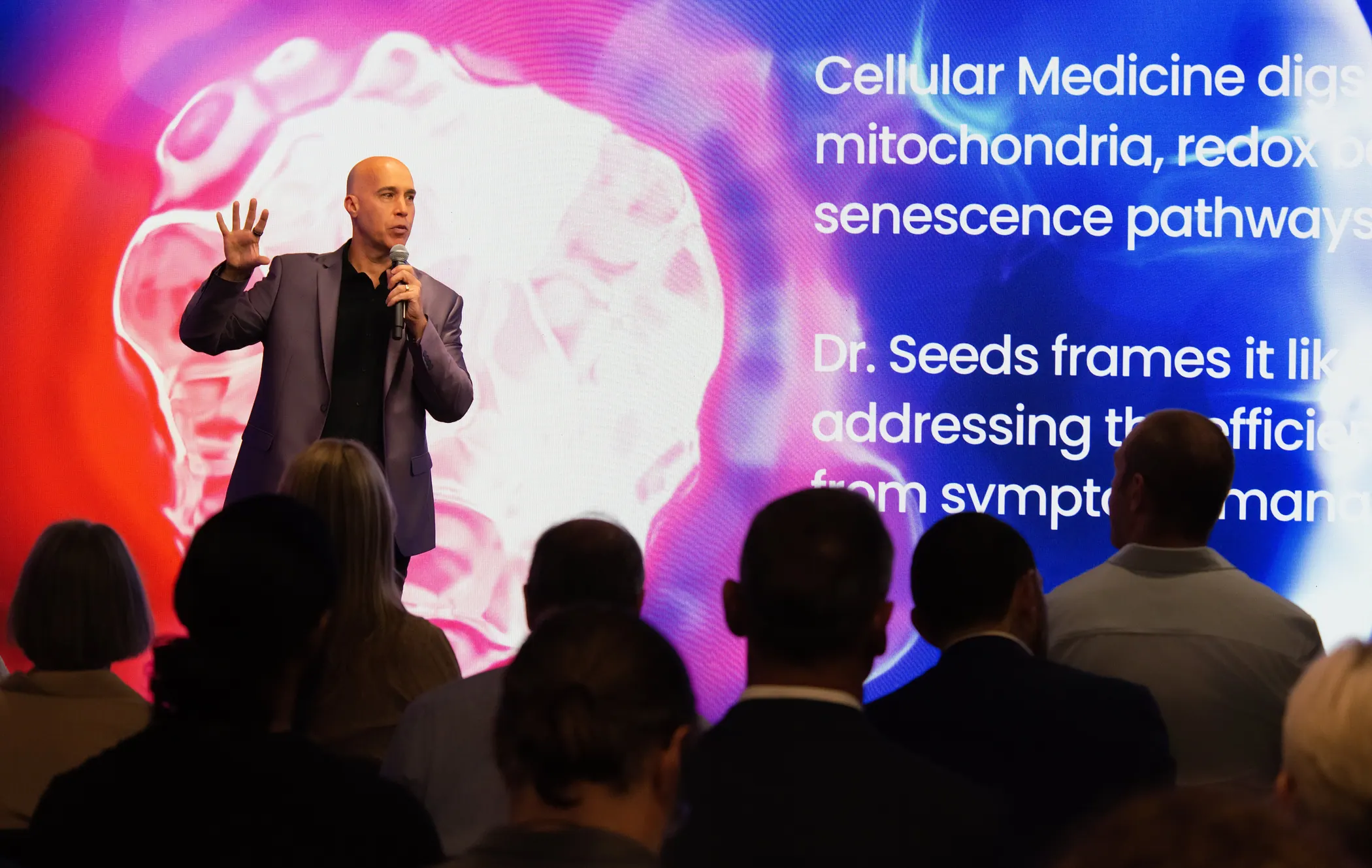 Elias Arjan mid-gesture on stage in front of a vivid cellular-medicine slide